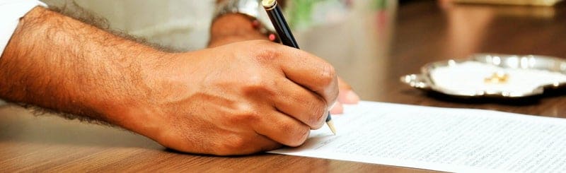Mano firmando un documento con una pluma sobre una mesa de madera